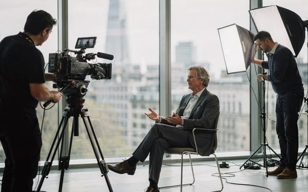 A two-person film crew shooting a seated business executive in a bright modern office, one operating a cinema camera on a tripod while the other adjusts a softbox light