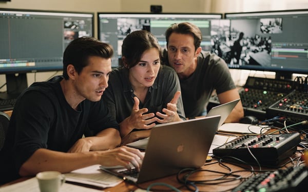 Two video editors and a producer reviewing older footage on a laptop in a busy edit suite surrounded by monitors