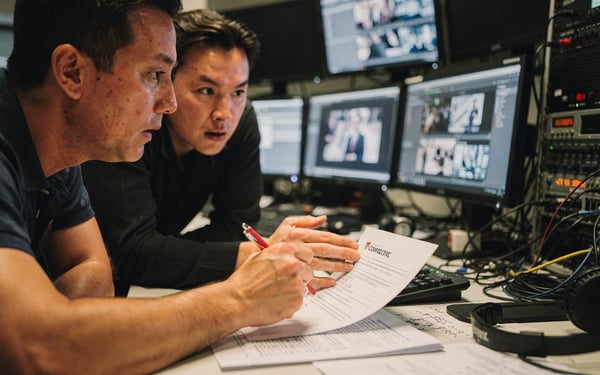 Two colleagues in an edit suite reviewing footage on monitors and marking up a printed script