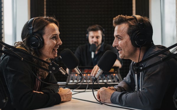 Two podcast presenters in headphones talking into microphones in a recording booth while a producer adjusts levels nearby
