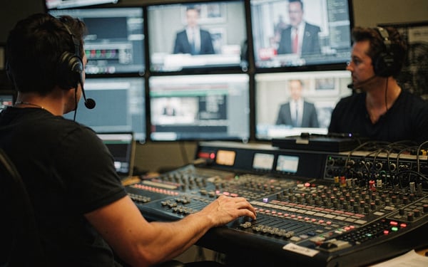 Two production crew members in a broadcast control gallery watching a live presenter feed across vision-mixing desks and monitors, one adjusting a fader