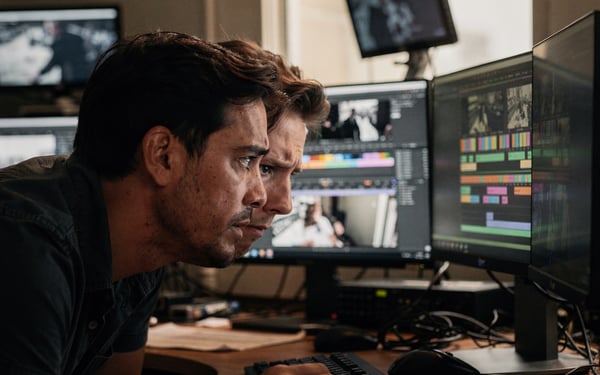 Two men lean in close to study editing software on dual monitors in a dim edit suite, frowning at the footage