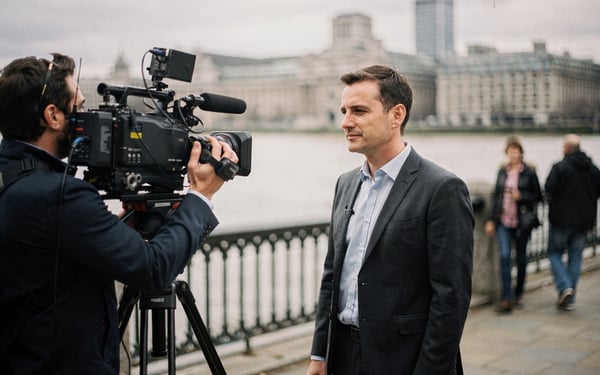 A camera operator films a presenter in a grey suit on the South Bank promenade beside the Thames, with the London skyline and people walking behind