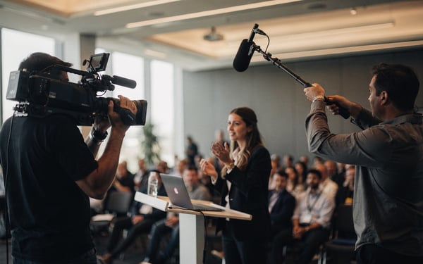 A video crew films a businesswoman giving a keynote on a conference stage in front of a seated audience
